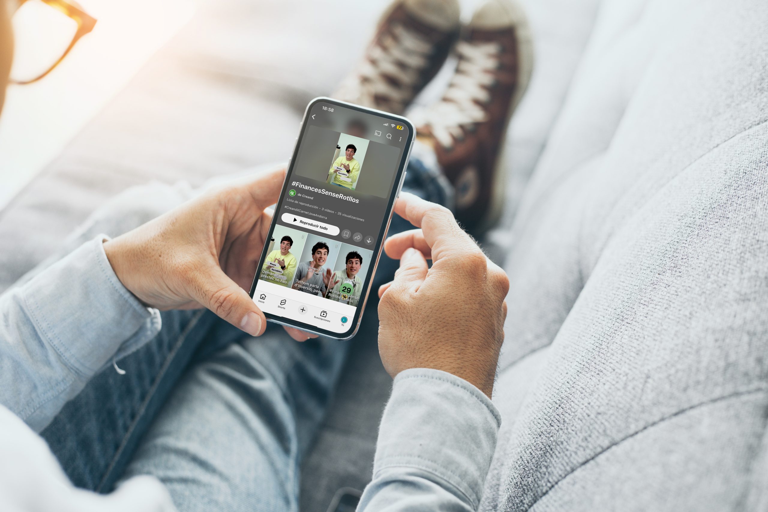 Persona asseguda en un sofà, sostenint un mòbil que mostra un perfil de xarxa social amb el títol #FinancesSenseRotllos i diverses miniatures de vídeo.       
