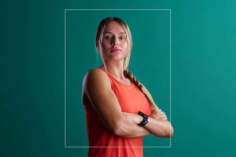 Retrat d'una dona en roba esportiva taronja, amb els braços plegats i un rellotge intel·ligent, sobre un fons verd fosc.