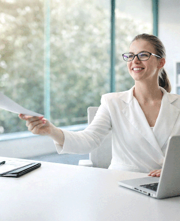 Asesora de seguros sonriendo, con gafas y chaqueta blanca, sentada en una mesa de oficina, ofreciendo un documento a un cliente con un ordenador portátil delante y luz natural de fondo. 