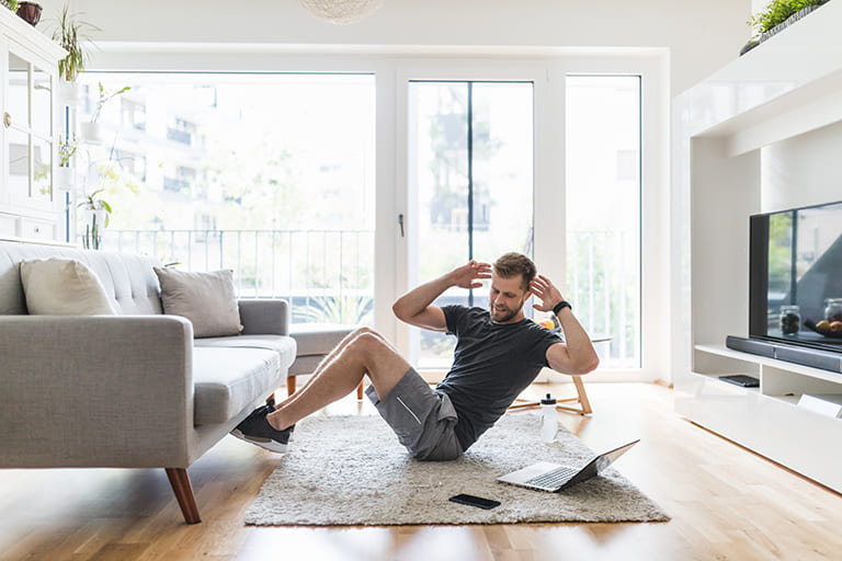 A young man doing sit-ups in his living room, following a workout routine on a laptop.