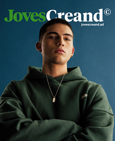 People 9 Young man from eighteen to thirty years old wearing a dark green hooded sweatshirt and a silver collar looking at the camera with a defiant expression on a blue studio background and under the JovesCreand logo